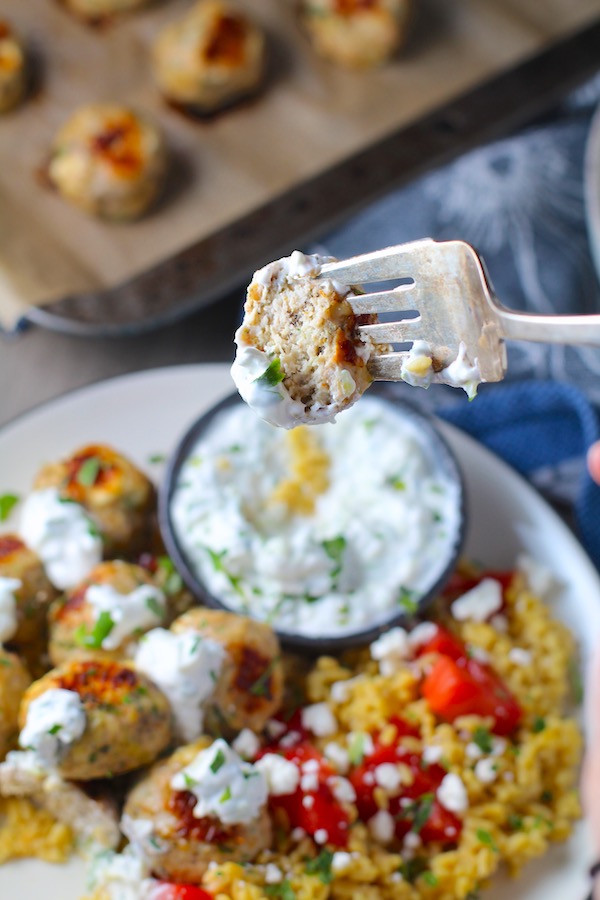 Ground Chicken Greek Meatball on fork dipped in Tzatziki sauce over rice. Baked meatballs in back. These meatballs have garlic, feta, parsley, and oregano