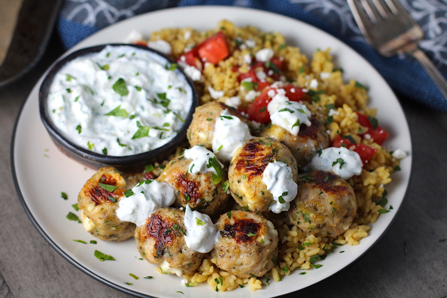 Ground Chicken Greek Meatballs on a plate with Tzatziki sauce over rice. Baked meatballs in back. These meatballs have garlic, feta, parsley, and oregano. 