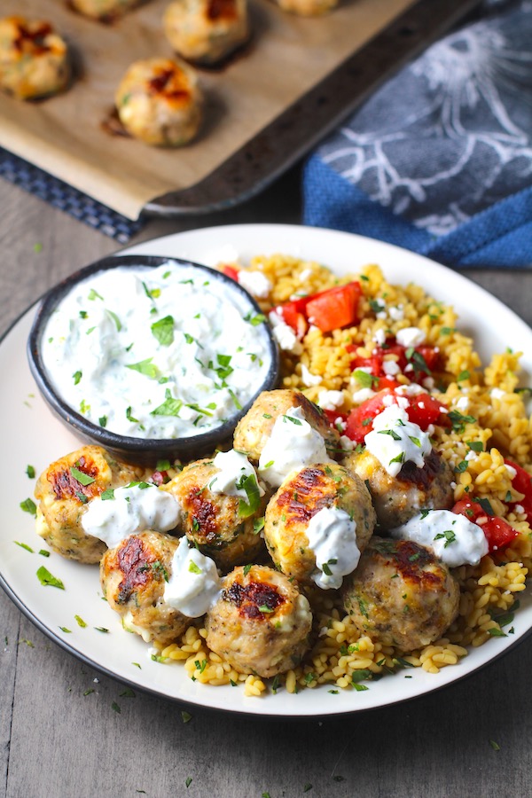 Ground Chicken Greek Meatballs on a plate with Tzatziki sauce over rice. Baked meatballs in back. These meatballs have garlic, feta, parsley, and oregano