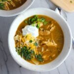 Creamy Chicken Enchilada Soup in a bowl topped with cheese, sour cream, and cilantro! It's creamy, hearty, slightly spicy with green chiles and smokey seasonings that bring so much depth.  Corn tortilla strips act like the noodles in this soup!