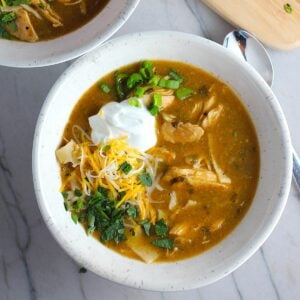 Creamy Chicken Enchilada Soup in a bowl topped with cheese, sour cream, and cilantro! It's creamy, hearty, slightly spicy with green chiles and smokey seasonings that bring so much depth. Corn tortilla strips act like the noodles in this soup!