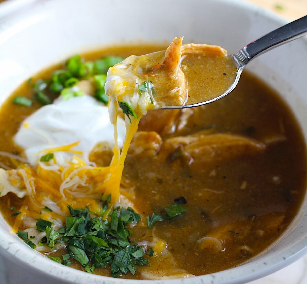 Spoon with Creamy Green Chicken Enchilada Soup in a bowl topped with cheese, sour cream, and cilantro! It's creamy, hearty, slightly spicy with green chiles and smokey seasonings that bring so much depth. Corn tortilla strips act like the noodles in this soup!