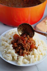 Ladling Chili over Mac and Cheese on a plate. This Recipe has creamy and cheesy pasta topped with smokey, tangy, savory beef chili! #macandcheese #chilirecipes