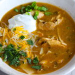 Creamy Chicken Green Enchilada Soup in a bowl topped with cheese, sour cream, and cilantro! It's creamy, hearty, slightly spicy with green chiles and smokey seasonings that bring so much depth.  Corn tortilla strips act like the noodles in this soup!