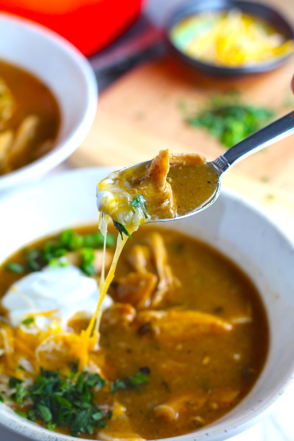 Spoon with Creamy Chicken Enchilada Soup in a bowl topped with cheese, sour cream, and cilantro! It's creamy, hearty, slightly spicy with green chiles and smokey seasonings that bring so much depth.  Corn tortilla strips act like the noodles in this soup!