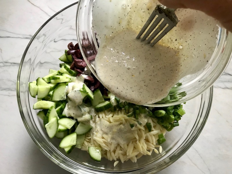 Pouring dressing over Greek Orzo Salad with Kalamata Olives, Cucumbers, and Feta Cheese.  It's mixed with a creamy lemon dressing that's bright and light!