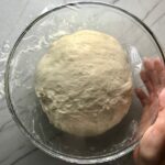 Homemade pizza dough in a clear bowl covered with plastic wrap and hand on side for a BBQ Chicken Pizza Recipe