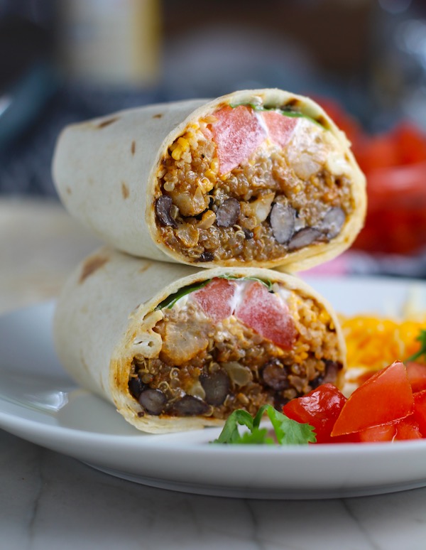 Quinoa Black Bean Burritos cut in half and stacked on plate. Packed with protein and fiber, but also cheese, tomato, cilantro, and sour cream!  The vegetarian 'meat' is taco-seasoned quinoa, black beans, and cauliflower.