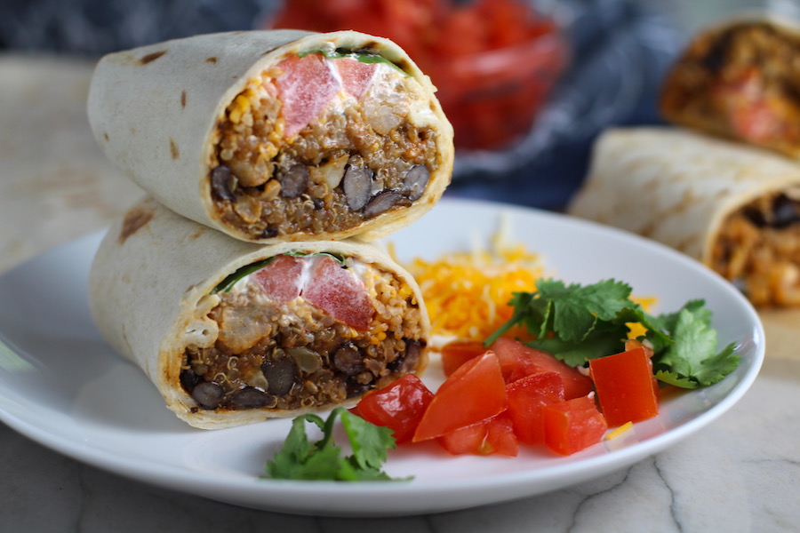 Quinoa Black Bean Burritos cut in half and stacked on plate. Packed with protein and fiber, but also cheese, tomato, cilantro, and sour cream!  The vegetarian 'meat' is taco-seasoned quinoa, black beans, and cauliflower.