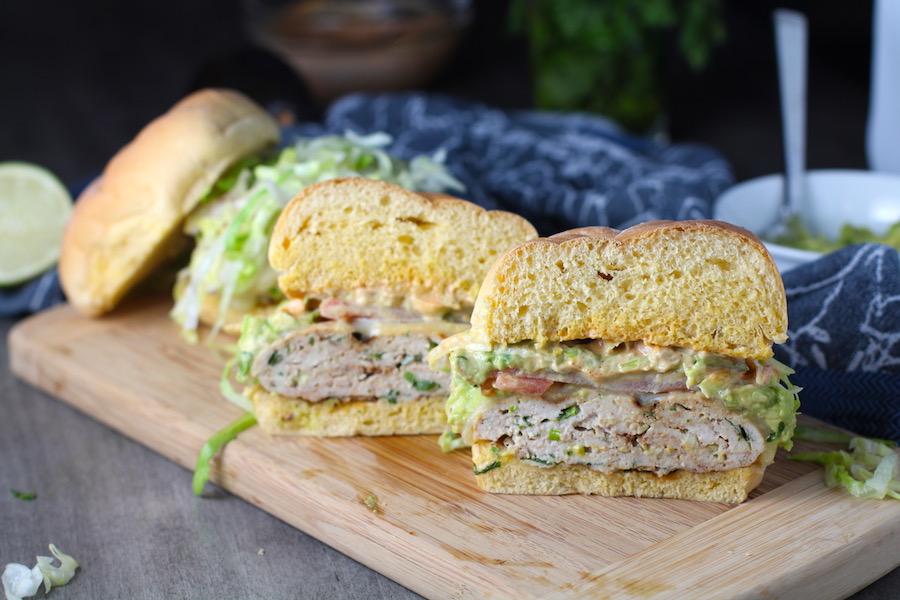 Taco Chicken Burger cut in half on wood plate. The burger has smokey taco seasonings, cilantro, and scallions. Then on top are melty cheese, guacamole, lettuce, tomato, and chipotle mayonnaise!