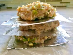 Raw chicken patties stacked between aluminum foil pieces for a Taco Chicken Burger recipe. The burger has smokey taco seasonings, cilantro, and scallions. Then on top are melty cheese, guacamole, lettuce, tomato, and chipotle mayonnaise!