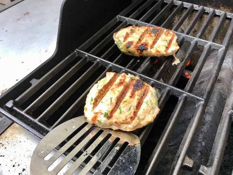 Grilled chicken burger on gas grill for a Taco Chicken Burger recipe. The burger has smokey taco seasonings, cilantro, and scallions. Then on top are melty cheese, guacamole, lettuce, tomato, and chipotle mayonnaise!