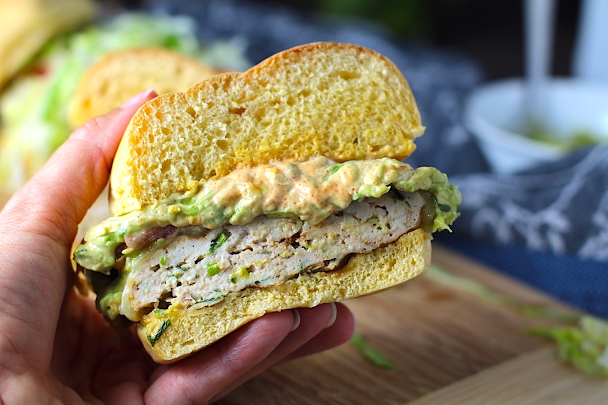 Hand holding half of a Turkey Taco Burger with smokey taco seasonings, cilantro, and scallions. Then on top are melty cheese, guacamole, lettuce, tomato, and chipotle mayonnaise!