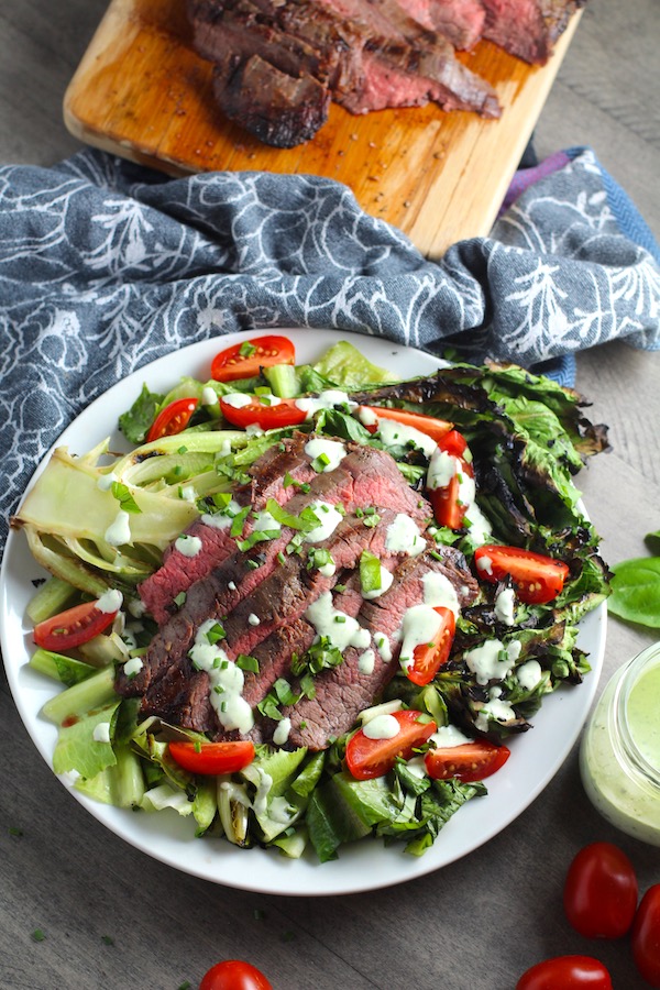 Grilled Flank Steak Salad on a plate with towel behind it.  It has garlic-rubbed flank steak over grilled romaine, bright tomatoes with basil yogurt dressing drizzled over top.
