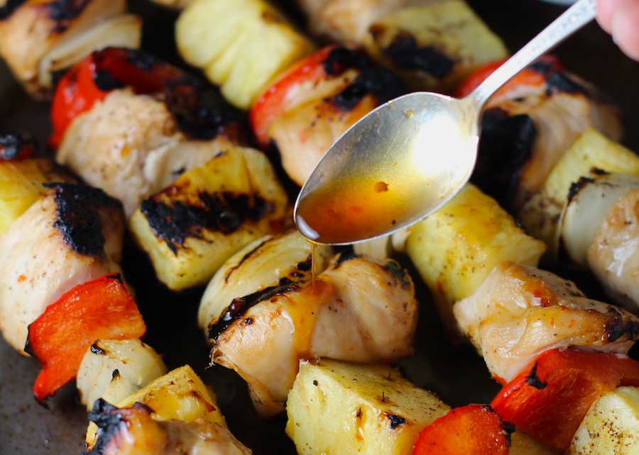 Spoon drizzling Sweet and Sour sauce over Chicken Kabobs with pineapple, onion, and red pepper on a pan.