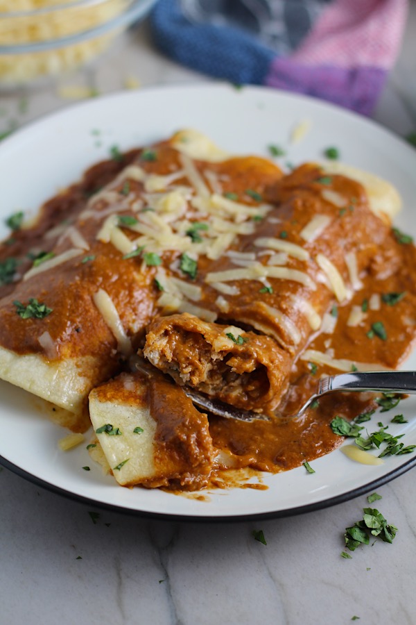 Fork with bite of Chicken Mole Enchiladas, on a plate with cheese and cilantro on top.  They are easy to make and delicious!  The Mole sauce has unsweetened chocolate, tomatoes, green chilis, garlic, adobo sauce, and spices blend harmoniously!  