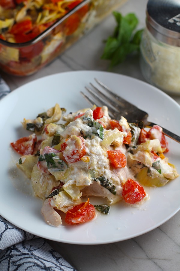 Chicken Artichoke Bake with Tomatoes and Goat Cheese on a plate with fork and casserole in back.   It's a creamy, tangy, sweet, savory, mouth watering easy dinner that you can prep ahead!