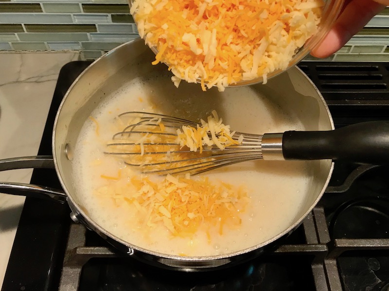 Shredded cheddar cheese being added to thickened sauce for Broccoli Quinoa Casserole recipe.