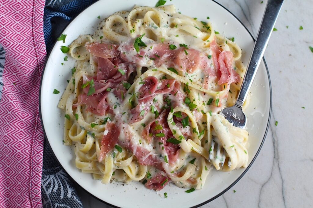 Fettuccine Lower Fat Alfredo Sauce Recipe with Prosciutto on a plate with fork scooping pasta. It's a creamy but lighter recipe without the heavy cream or butter, but still delicious!