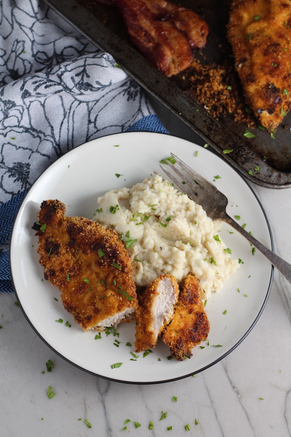 Crispy Baked Chicken Breast Cutlets with Bacon Crumbs and Honey cooked on a plate with part of it sliced and mashed cauliflower and fork on plate.  Sheet pan in background.