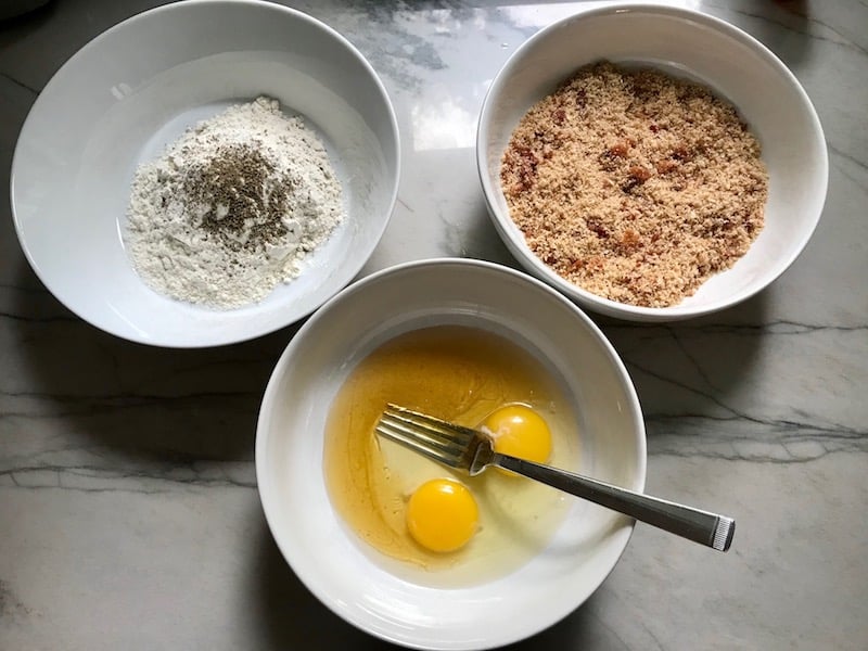 3 bowls on counter for Oven Fried Chicken Breast. One bowl has flour, another has honey and eggs, and the last has breadcrumbs. The chicken is coated with Bacon Crumbs and Honey and it's healthier because it's baked, not fried, but still gives you all of the crispy, crunchy goodness!  It also gives you sweet and salty crunch with the addition of honey and bacon!