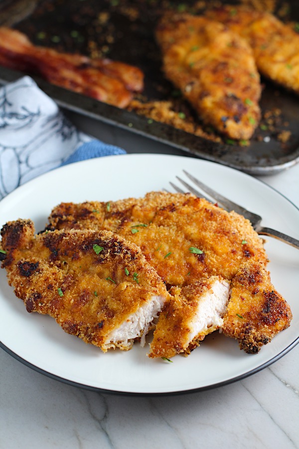 Crispy Baked Chicken Breast Cutlets with Bacon Crumbs and Honey cooked on a plate with part of it sliced and fork on plate.  Sheet pan in background.