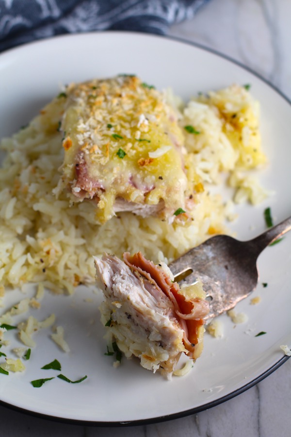 Piece of One Pot Chicken Cordon Bleu Bake on a plate with fork holding a bite.  On the bottom you get flavorful rice topped with chicken, salty ham, nutty and melty swiss cheese, and a crunchy breadcrumb topping!