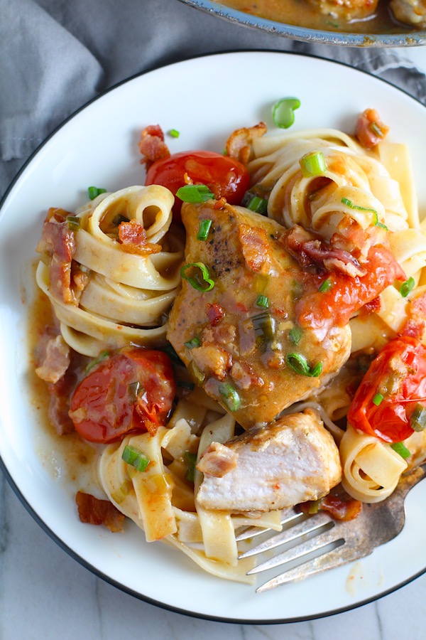 Smothered Chicken with Bacon, and Tomatoes and fettuccine on a plate. The chicken is smothered in a thickened sauce infused with smokey and salty bacon flavor.  Cherry tomatoes give a sweet and tangy pop.