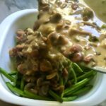 Chorizo and mushroom sauce being poured over green beans in casserole dish for Creamy Green Beans and Mushrooms with Chorizo in a serving bowl are a creamy and savory side dish.