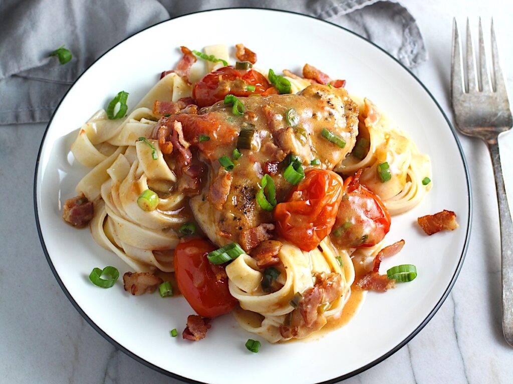 Smothered Chicken with Bacon, and Tomatoes and fettuccine on a plate with pan in background. The chicken is smothered in a thickened sauce infused with smokey and salty bacon flavor.  Cherry tomatoes give a sweet and tangy pop.