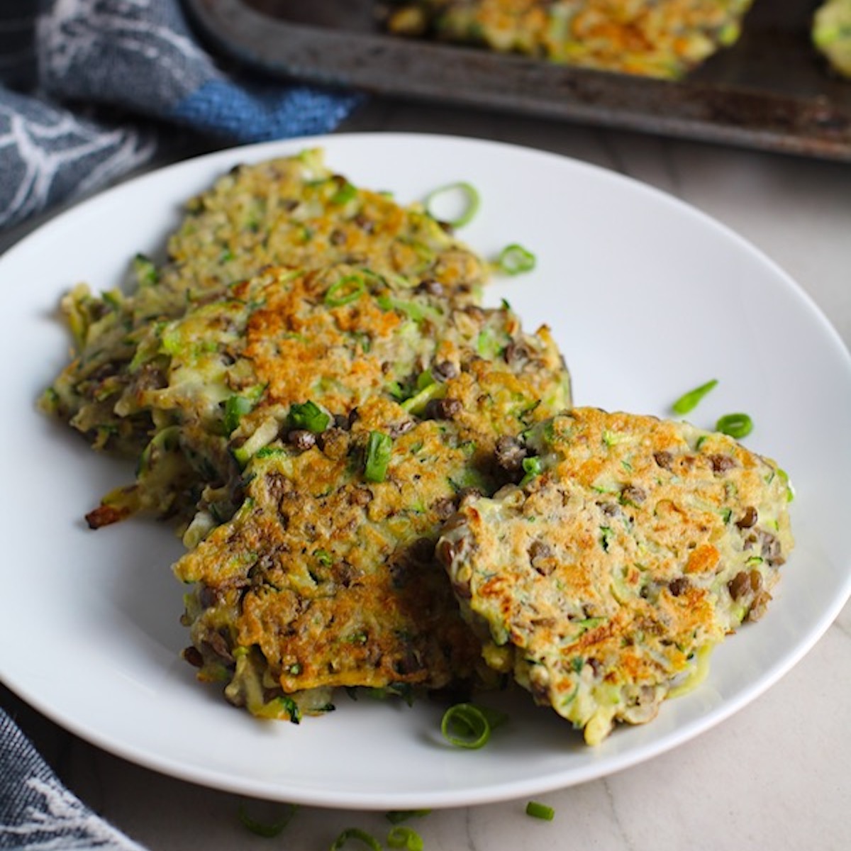 Zucchini Lentil Fritters on a plate with more on a pan in the background. They have a crispy outside and soft inside and are perfect for an easy dinner!  The flavors are simple but perfect with parmesan, oregano, scallions, lentils, and zucchini.