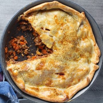 Flaky Empanada Pie with Ground Beef Recipe in a skillet with a piece out on counter. It has a perfect flaky, buttery pastry on the outside with a savory, smokey, salty ground beef filling.  The best part is that everything cooks in one skillet!