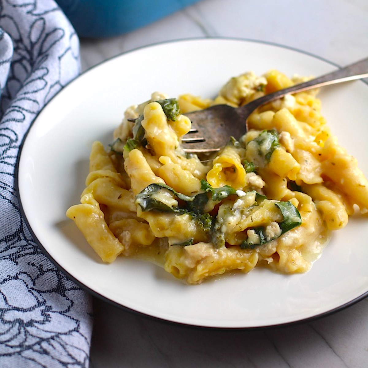 Greek Ground Chicken Feta Pasta Casserole on a plate with fork and casserole in background. It has lean ground chicken with garlic, oregano, parmesan, feta and Kale. 