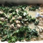 Fork pressing down dry pasta and kale into liquid with ground chicken and broth for Greek Ground Chicken Feta Pasta Casserole. It has lean ground chicken with garlic, oregano, parmesan, feta and Kale. 