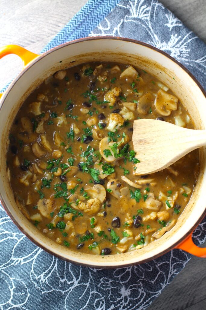 Creamy Cajun Cauliflower and Beans in apot with wood spoon.  The mild cajun flavors combined with  healthy garbanzo beans, black beans, cauliflower, and mushrooms make this a hearty, creamy, and delicious dish
