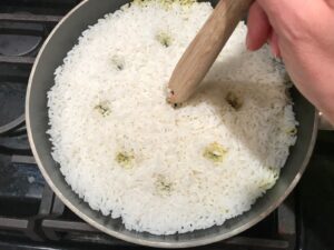 Spoon handle poking holes into cooked rice in pan for Saffron Butter Crispy Rice. Saffron brings a floral note and beautiful yellow-orange color.  Added to the butter is what gives this crispy rice the buttery, crispy bottom.