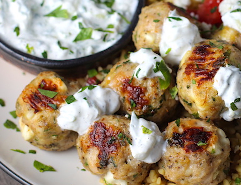 Ground Chicken Greek Meatballs on a plate with Tzatziki sauce over rice. Baked meatballs in back. These meatballs have garlic, feta, parsley, and oregano. 