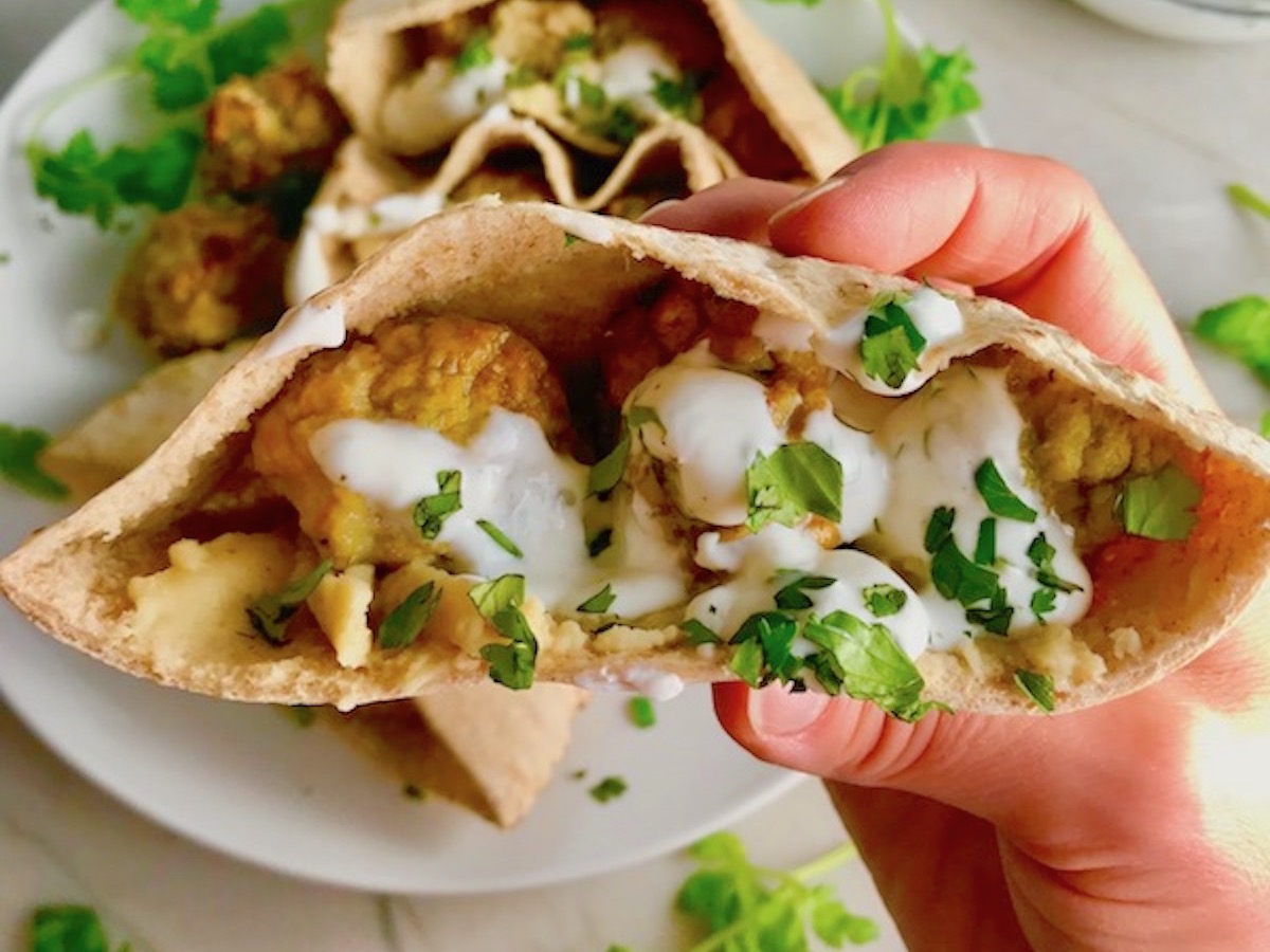 Hand holding Moroccan Pita Bread Sandwiches Recipe on a plate. They are filled with homemade hummus, moist turkey meatballs made with warm Moroccan spices like ginger, turmeric, and coriander, and then topped with creamy, fresh and bright lemon yogurt sauce.