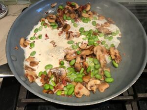 Cream, mushrooms, edamame, and Bacon bits cooking in pan for Pasta with Ricotta, Bacon, Mushroom Sauce on stove. It's creamy, rich, decadent, comforting, and downright delicious!