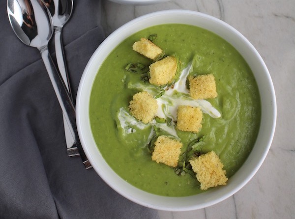 Zesty Pea Soup with Spring Peas, Jalapeño, and Mint in a bowl with crema and croutons on top and spoon on the side.