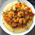 Cajun Shrimp and Grits recipe on a plate. It has seared shrimp in a creamy smoky cajun sauce with chicken andouille sausage and pimiento.  Served over creamy, cheesy grits.