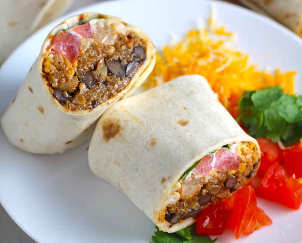 Burritos with Black Beans and Quinoa recipe cut in half and stacked on plate. Packed with protein and fiber, but also cheese, tomato, cilantro, and sour cream! The vegetarian 'meat' is taco-seasoned quinoa, black beans, and cauliflower.