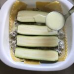 Ladle pouring white sauce over zucchini slices on top of beef and lasagna noodles in dish for White Lasagna Recipe with Zucchini and Ground Beef
