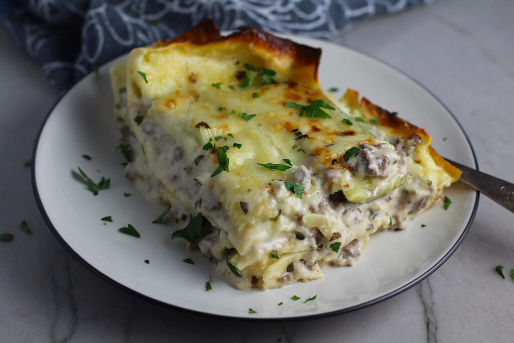 Piece of White Lasagna Recipe with Zucchini and Ground Beef with browned melted cheese on top on a plate with parsley. It's a make-ahead family dinner! It's creamy, hearty, and delicious with layers of noodles, zucchini, ground beef, creamy cheese, and a silky white sauce. 