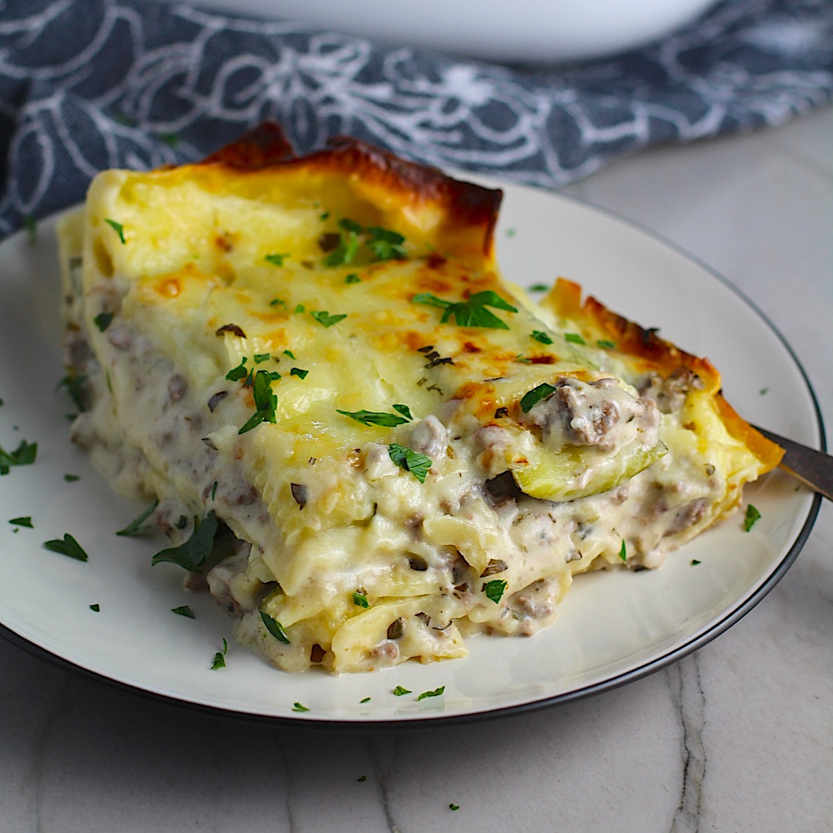 Piece of Lasagna with Cream Cheese, Beef, Zucchini with browned melted cheese on top on a plate with parsley. It's a make-ahead family dinner! It's creamy, hearty, and delicious with layers of noodles, zucchini, ground beef, creamy cheese, and a silky white sauce. 