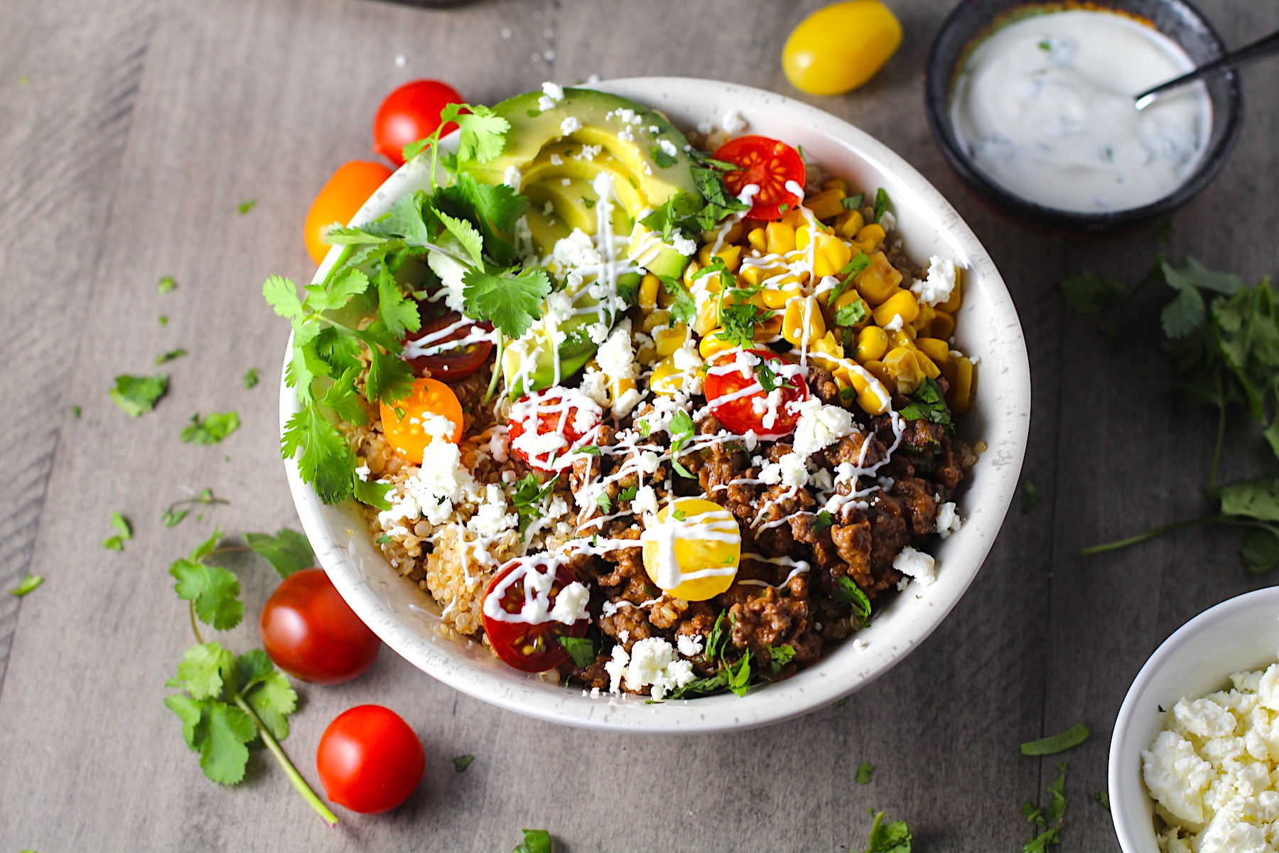 Taco Quinoa Bowl with Ground Beef, corn, cotija cheese, tomatoes, fresh cilantro, and cilantro lime crema drizzled on top. Bowl of crema, bowl of cotija cheese, tomatoes and cilantro on the table.