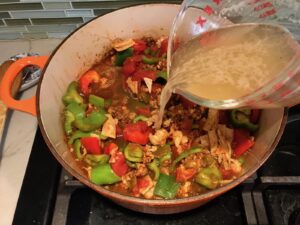 Pouring broth into pot with ground chicken cooking, peppers, and tomatoes for Tex Mex Unstuffed Peppers with Ground Chicken.  Juicy Sweet Bell Peppers are chopped up and cooked with the Tex Mex style ground chicken, tomatoes, Mexican seasonings, cheese, rice and corn tortillas.