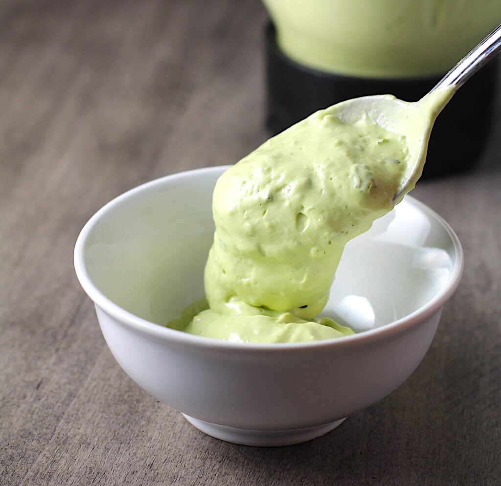 Spoon pouring Avocado Crema Recipe to a bowl on table with blender in back. It goes on everything! It's a creamy, tangy, rich and delicious sauce that's made in just minutes in the blender!