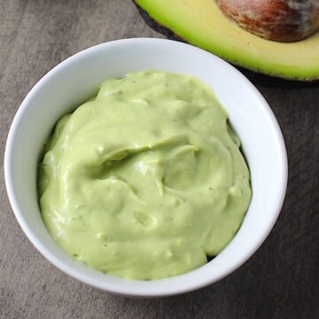 Easy Avocado Crema Recipe in a bowl on table with a cut open avocado with pit. It goes on everything! It's a creamy, tangy, rich and delicious sauce that's made in just minutes in the blender!