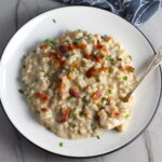 Velvety Barley Recipe with Bacon and Gruyere Cheese on plate with crispy bacon bits and diced chives on top and spoon with scoop on side of plate. It's creamy, rich, nutty, smokey, hearty, and utterly delicious.